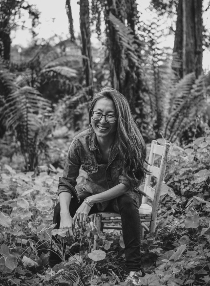 [Black and white photo of a korean woman smiling with long hair and glasses, sitting on a wooden chair with plants and trees all around her.]
