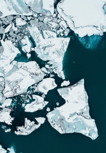 Photograph of a melting iceberg taken from above. White pieces of ice of varying sizes of all different shapes float over dark blue water. The ice is white, with shades of grey as well and some of it is submerged under water, creating a ghostly white-blue. There is a main large piece of ice that is towards the center of the frame, in a triangular-is shape and most of the other ice descends from the left upper corner. Some are clustered close together, while others float farther out. The photo is beautiful and almost looks surreal and like a piece of abstract art.